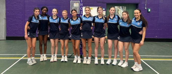 10 girls in sports kit line up for team photo in a sports hall in front of a netball hoop